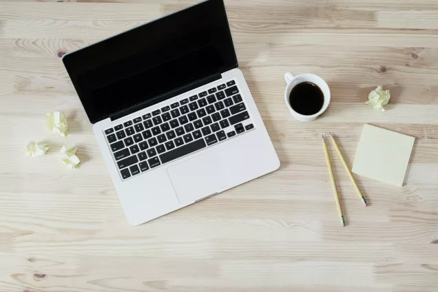 Bild på en laptop, en kopp kaffe, post it-lappar och pennor, sett ovanifrån. Foto.